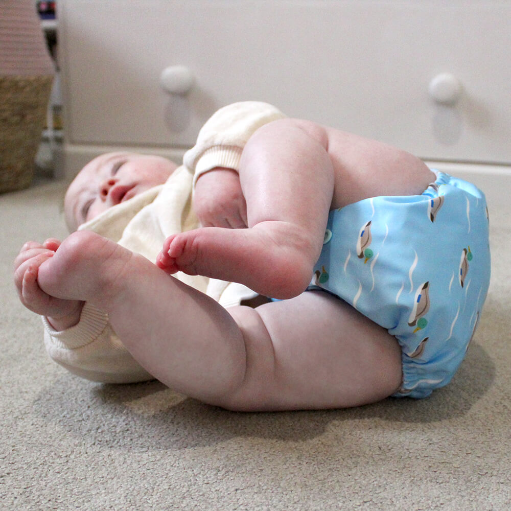 A baby wearing a blue cloth nappy with ducks, lying on the floor holding his toes.